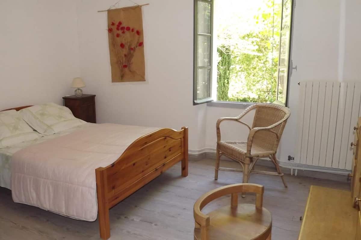 A bedroom with a wooden bed, a wicker chair, a small table, and a window with a view of greenery.