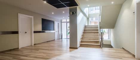 Un pasillo amplio con piso de madera, una escalera y una puerta con letrero de salida de emergencia.