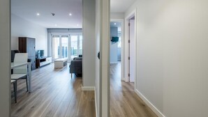 Eine moderne Wohnung mit Holzboden, einem Essbereich mit weißem Tisch und Stühlen, einem Wohnzimmer mit Sofa und Fernseher sowie Blick auf die Stadt durch große Fenster.