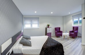 Un dormitorio moderno con una cama, dos sillones morados, una mesita y una ventana con vistas.
