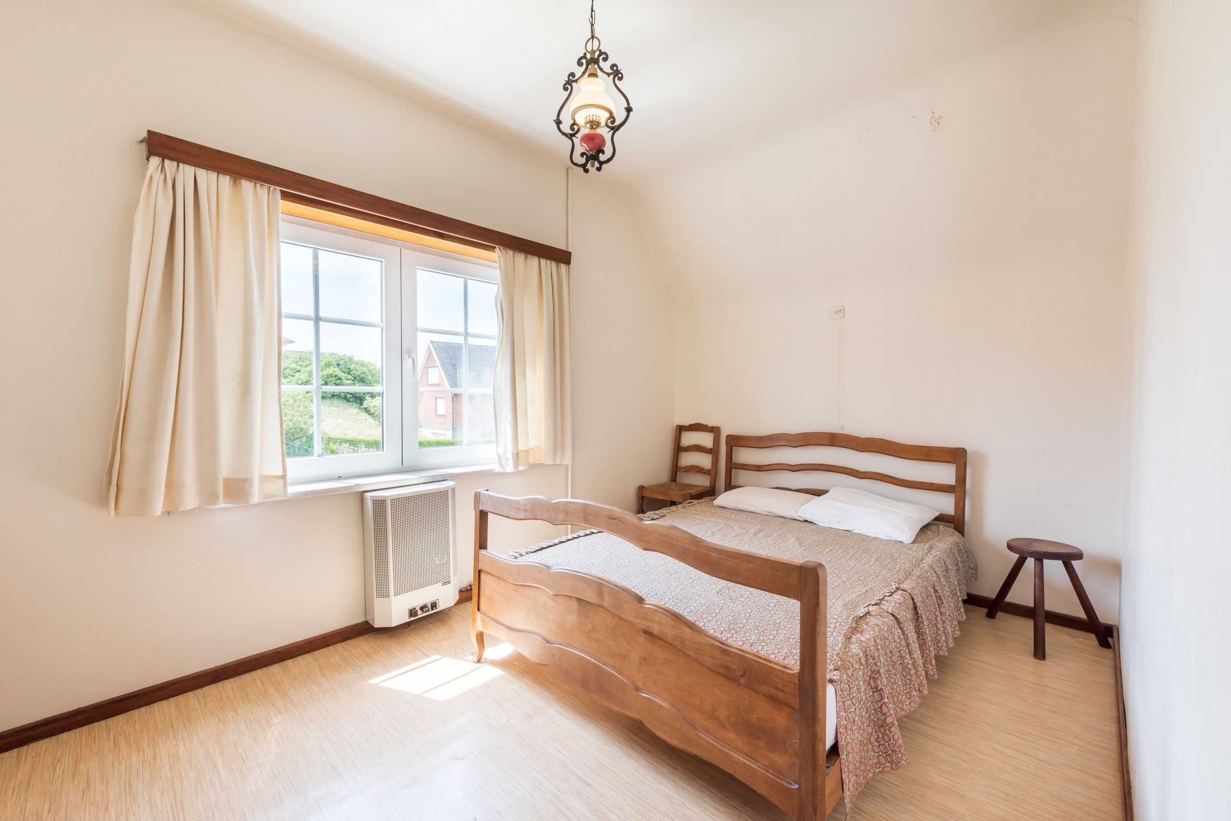 Quarto com cama de madeira, radiador, janela com cortinas e luminária suspensa.