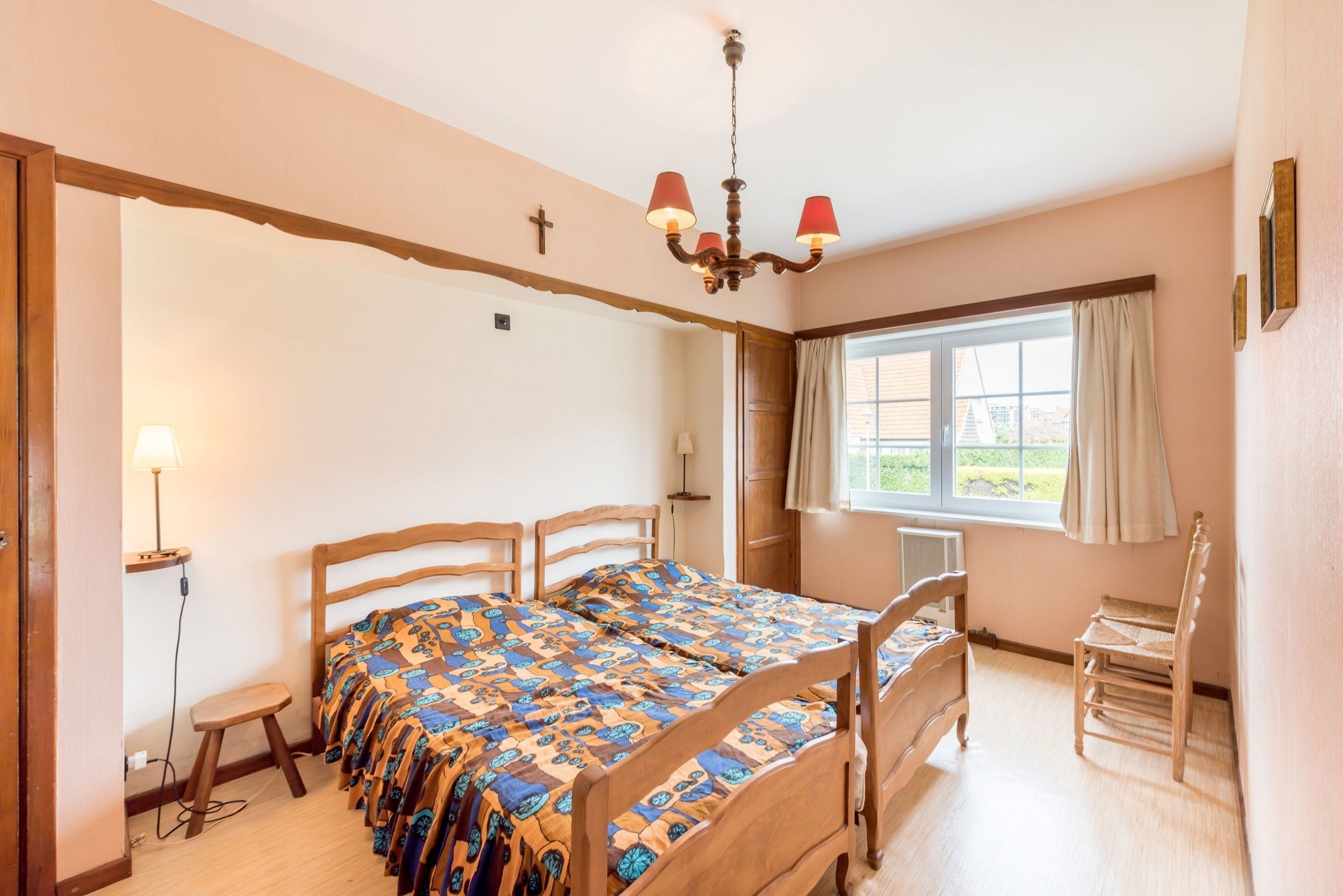 A bedroom with a wooden bed, a nightstand, a chair, a window with curtains, and a chandelier.