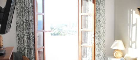 A bedroom with a bed, a chair, a desk, a lamp, and an open door leading to a balcony.