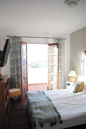 Ein Schlafzimmer mit Bett, Stuhl, Schreibtisch, Lampe und einer offenen Tür, die auf einen Balkon führt.