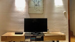 Un téléviseur posé sur un meuble en bois, avec une télécommande, une petite table sur laquelle se trouve un livre, et une œuvre d’art accrochée au mur.