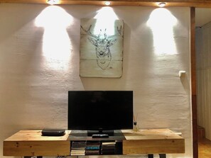 Un téléviseur posé sur un meuble en bois, avec une télécommande, une petite table sur laquelle se trouve un livre, et une œuvre d’art accrochée au mur.