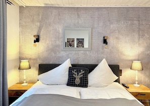 A bedroom with a concrete wall, a framed picture, two bedside lamps, and a deer-shaped pillow.