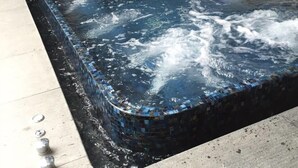 Un bain à remous rectangulaire, bordé de mosaïques, situé dans un espace extérieur doté d’un sol carrelé et d’une porte-fenêtre coulissante en arrière-plan.