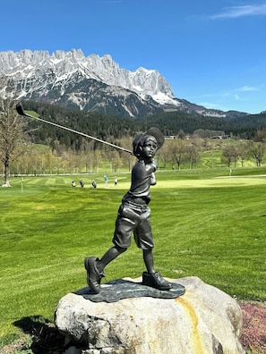 A statue of a person with a baseball bat on a rock, with a golf course and mountains in the background. - Cozy design apartment in the heart of Kitzbühel (Kitzbühel)