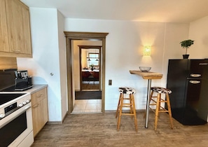 A kitchen with a stove, refrigerator, and stools. There is a doorway leading to another room.