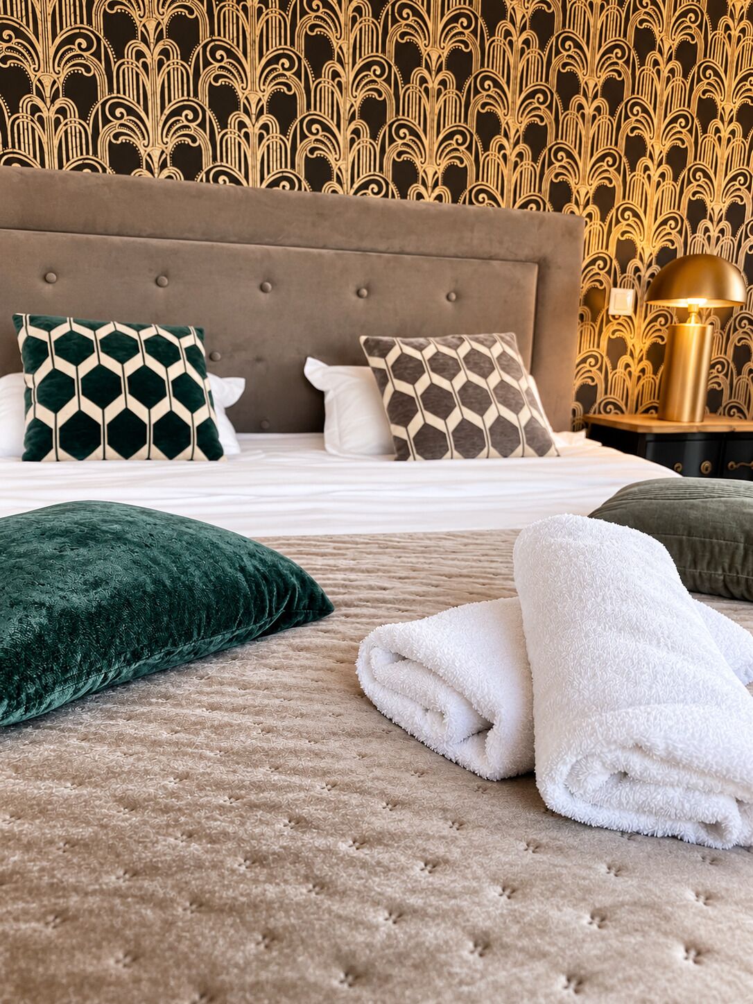 Un lit bien fait, avec une tête de lit capitonnée, deux coussins à motifs, un coussin vert et un ensemble de serviettes blanches pliées.
