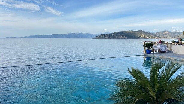 On aperçoit clairement une piscine, avec un palmier au premier plan, une terrasse en bois avec une table et des chaises, et un paysage montagneux en arrière-plan.