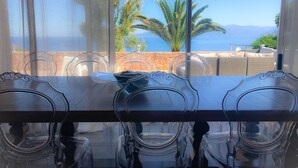 Un espace repas comprenant une table et des chaises, avec vue sur la mer et les montagnes, et un mur en pierre.