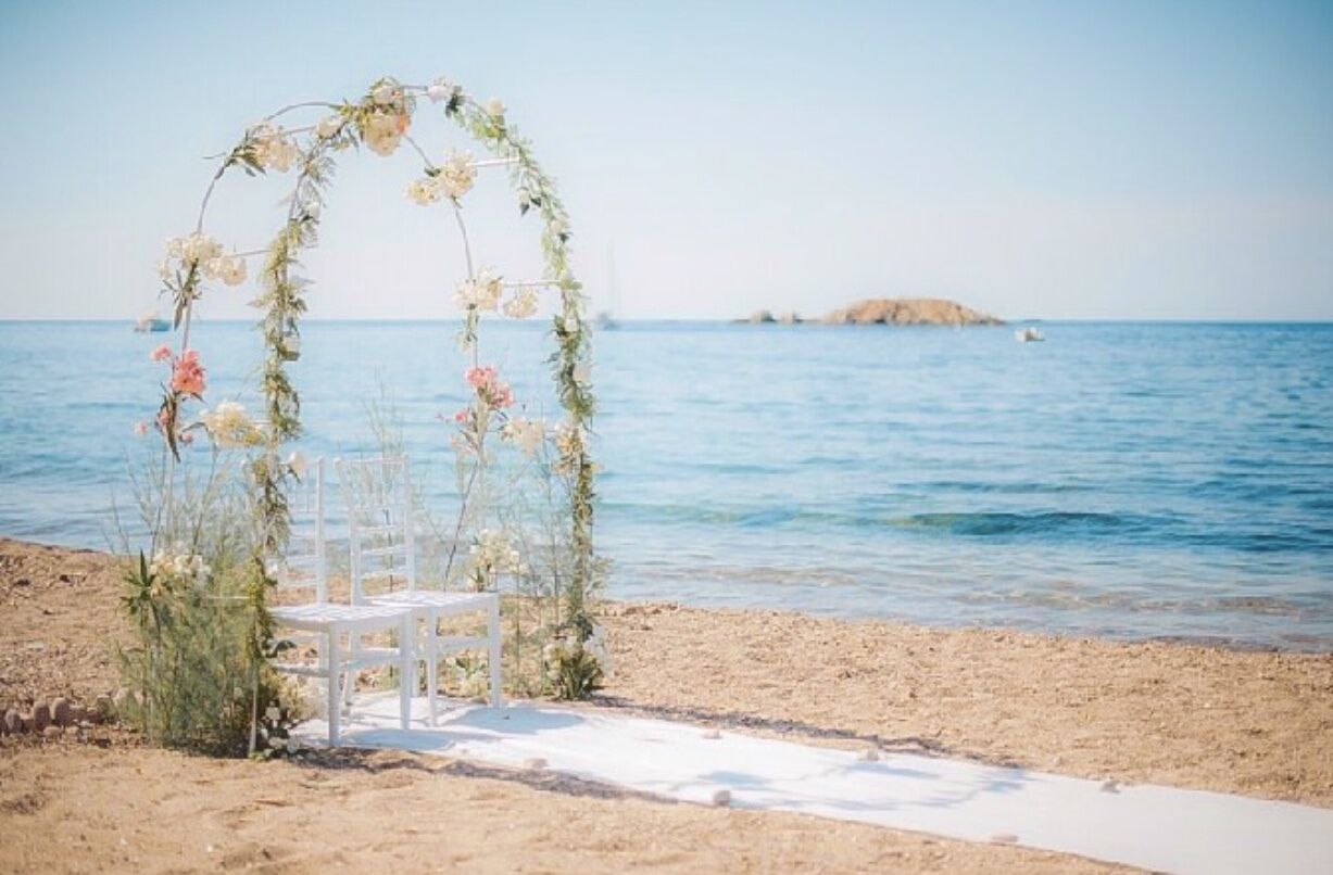 A beach wedding setup with a white arch adorned with flowers and a white chair.