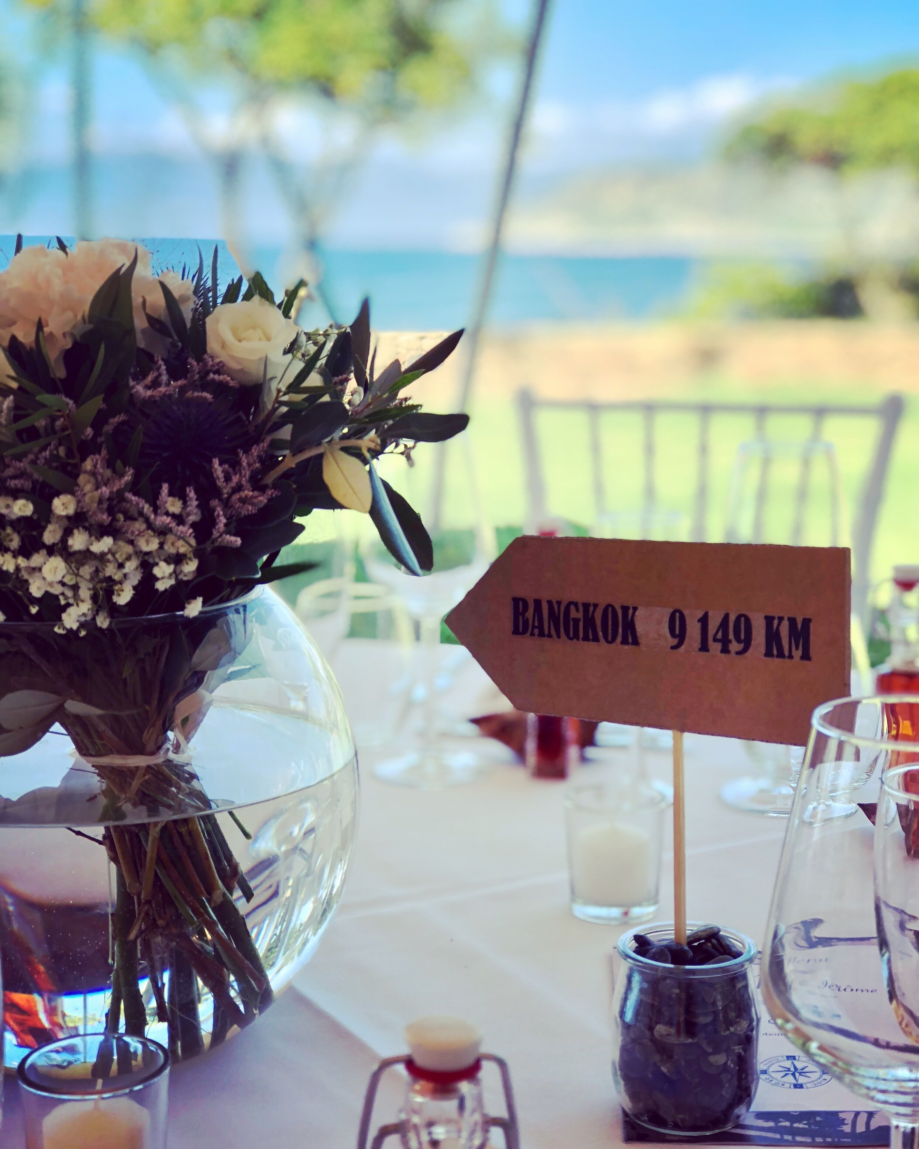 A table with a glass vase of flowers, a small sign with Bangkok 9149 KM, and a glass jar with black items.