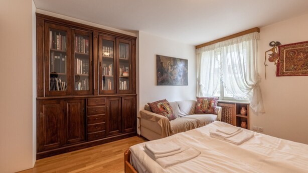 A bedroom with a large wooden wardrobe, a bed with neatly folded towels, a window with sheer curtains, and a wall-mounted artwork. - Casa Alverà (Cortina d´Ampezzo)