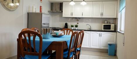 A dining area with a table set for four, wooden chairs, a clock on the wall, and a kitchen in the background.