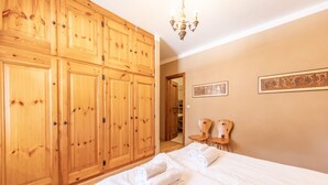 Une chambre à coucher avec une grande armoire en bois, un lit recouvert de draps blancs, deux chaises en bois et un lustre.