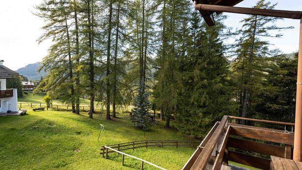A wooden deck with a view of a grassy area, trees, and a distant building. - Casa Pecol (Cortina d´Ampezzo)