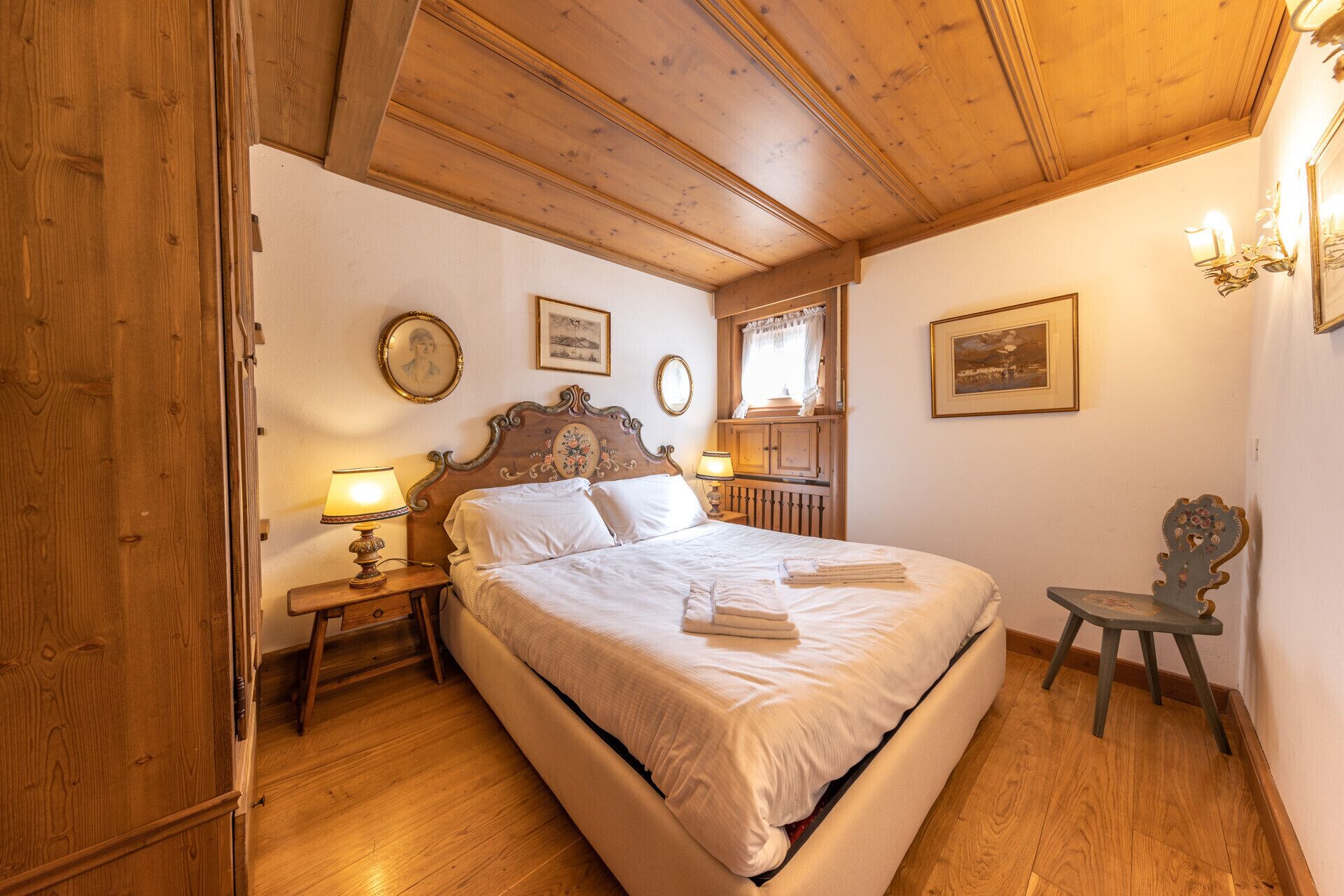 A bedroom with a wooden ceiling, a bed with white linens, two bedside tables with lamps, and framed pictures on the walls.