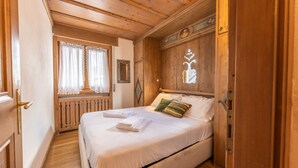 A wooden room with a bed, a window with curtains, and a wall with a decorative mirror and light fixture. - Casa Pecol (Cortina d´Ampezzo)