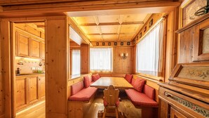 A wooden cabin interior with a central dining table, red cushioned benches, wooden cabinets, and a large window with curtains. - Casa Pecol (Cortina d´Ampezzo)