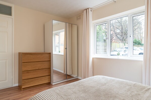 Une chambre avec une commode en bois, un lit recouvert d’une couverture blanche, un grand miroir et une fenêtre donnant sur des voitures garées et des arbres.