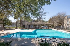 A swimming pool with a clear blue water, surrounded by a fenced area with trees and a building in the background.