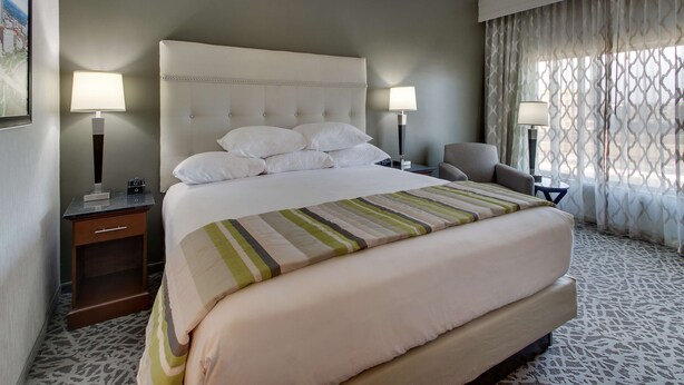 Quarto de hotel com uma cama grande, duas mesas de cabeceira com luminárias, uma cadeira e um tapete estampado.