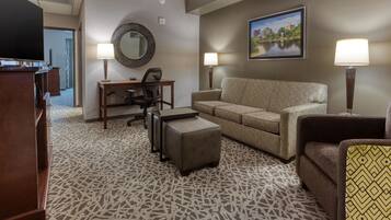 A hotel room with a sofa, armchair, desk, and ottoman. There is a television, a mirror, and two lamps. A painting is on the wall.