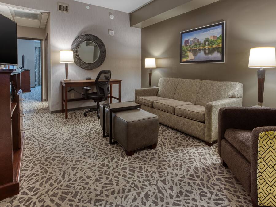 Ein Hotelzimmer mit Sofa, Sessel, Schreibtisch und Hocker. Es gibt einen Fernseher, einen Spiegel und zwei Lampen. Ein Gemälde hängt an der Wand.