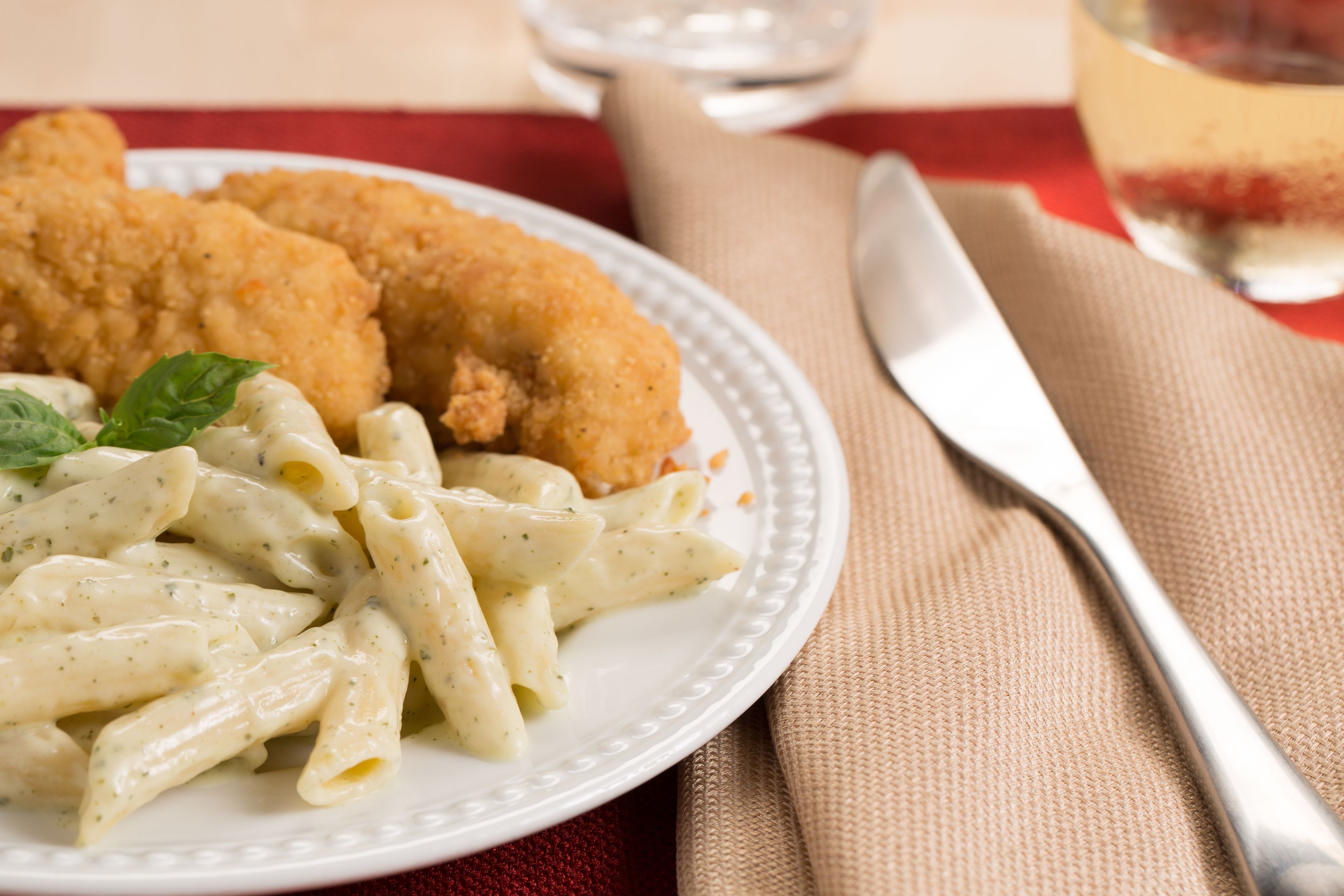 A plate of pasta with breaded cutlets and a garnish, accompanied by a fork and knife on a tablecloth.