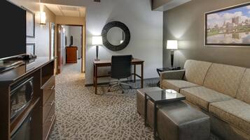A hotel room with a sofa, a TV, a desk with a chair, a mirror, and a patterned carpet.