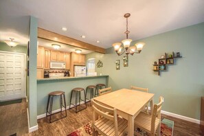 A dining area with a wooden table and chairs, a kitchen with wooden cabinets and a microwave, and a chandelier.