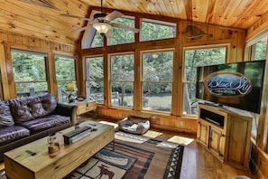 Une cabane aux murs en bois, dotée de grandes fenêtres, d’un ventilateur de plafond, d’une télévision, d’un canapé en cuir, d’une table basse et d’un tapis orné d’un motif d’élan.