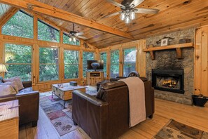 Interior de una cabaña con chimenea, sofá de cuero, muebles de madera y ventanas amplias.