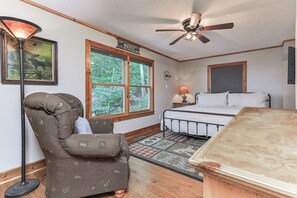 A bedroom with a bed, a chair, a standing lamp, a ceiling fan, and a window with a view of greenery. - Cartecay Canoe House | Ellijay, GA (Ellijay)