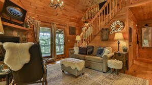 A cabin interior with a wooden staircase, a sofa, a chair with a fur throw, and a television on the wall. - Aska Retreat | Blue Ridge, GA (Cherry Log)