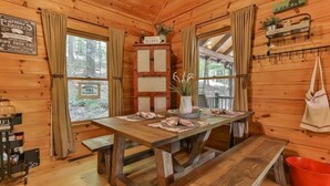 A wooden cabin interior with a wooden table, benches, a cabinet with signs, and a view of the forest through large windows. - Aska Retreat | Blue Ridge, GA (Cherry Log)