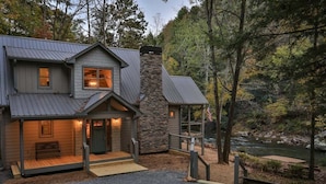 Una casa de dos pisos con una chimenea de piedra, un porche con un banco y vista a un río.
