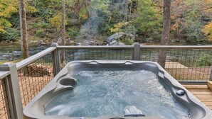 Un jacuzzi con panel de control digital, rodeado por una terraza de madera y baranda metálica, con vista a un río y zona boscosa.
