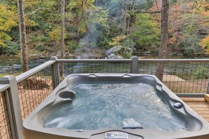 A hot tub with a digital control panel, surrounded by a wooden deck and a metal railing, overlooking a river and forested area.