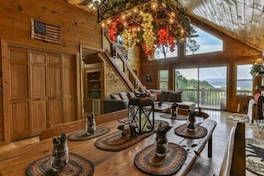 A wooden interior with a dining table set with placemats and candles, a staircase, and a view through large windows.