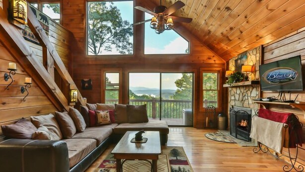 A cozy cabin interior with a wooden ceiling, a fireplace, a television, and a large window overlooking a scenic landscape. - Mountain Lake Escape | Ellijay, GA (Ellijay)