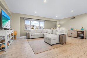 A modern living room with a sectional sofa, a flat-screen TV, and a large window with a view of buildings. - Beachfront 2BR condo w/ private beach access & views (Siesta Key)