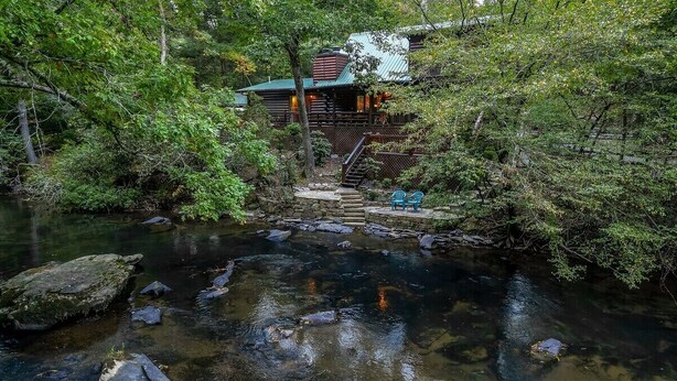 A cabin with a stone staircase leading to a deck, surrounded by dense forest and a clear stream. - River Run | Ellijay, GA (Ellijay)