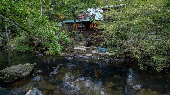 Une cabane avec un escalier en pierre menant à une terrasse, entourée d’une forêt dense et traversée par un ruisseau aux eaux claires.