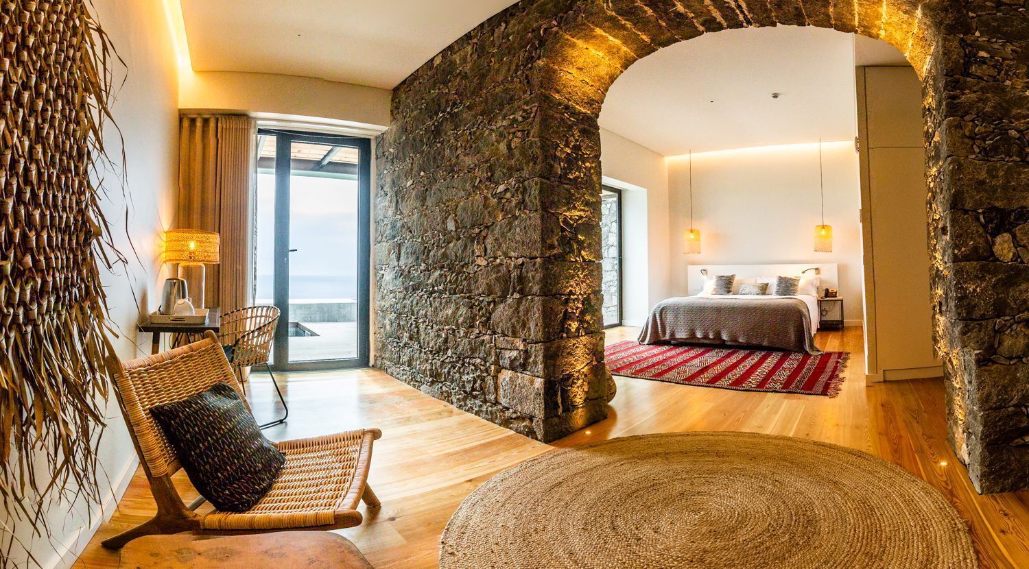 Ein Zimmer mit Steinwand, einem Bett, einem Korbstuhl, einem runden Teppich und Blick durch eine Glastür.