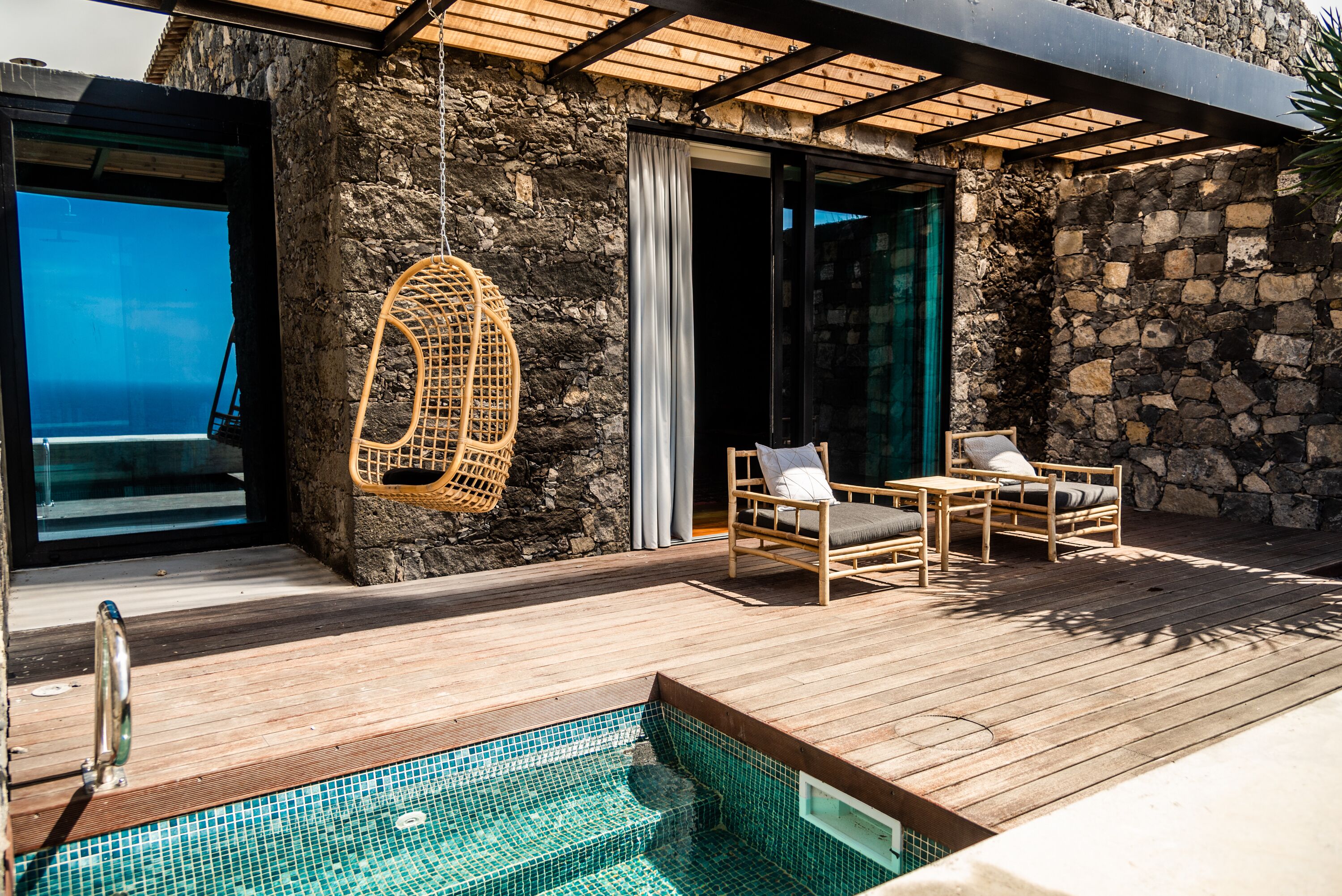Una terraza de madera con piscina, sillas de mimbre y un muro de piedra.
