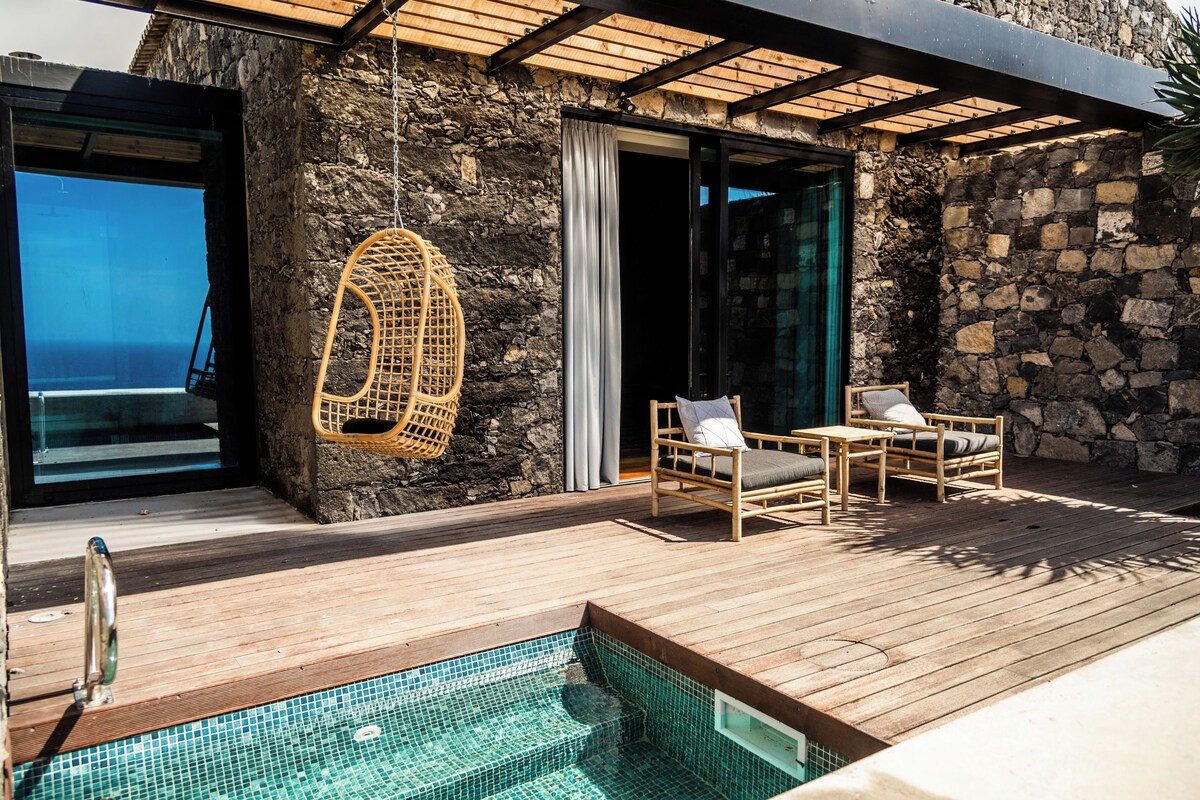 A wooden deck with a pool, wicker chairs, and a stone wall.